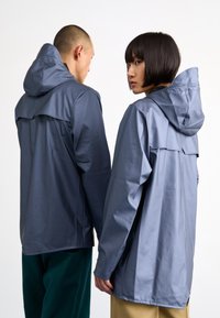 Vestes à capuche grises foncées, fabriquées en matériau lisse et imperméable. Équipées d'un empiècement au dos et de fentes latérales pour la ventilation. Coutures visibles.