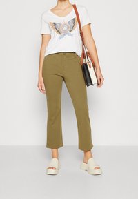 Vit T-shirt med en färgglad örn-grafik, olivgröna cropped byxor och beiga plattformssandaler. Brun handväska med svart och vit accents.