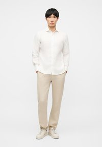 Άνδρας που φοράει λευκό πουκάμισο με μακριά μανίκια και κουμπιά, μπεζ παντελόνι ίσιας γραμμής και μπεζ αθλητικά παπούτσια, στέκεται με τα χέρια στις τσέπες.