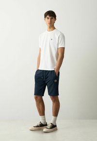 T-shirt bianca girocollo con un piccolo logo, abbinata a pantaloni corti blu navy con vita regolabile e tasche laterali. Sneakers marroni e calzini bianchi.