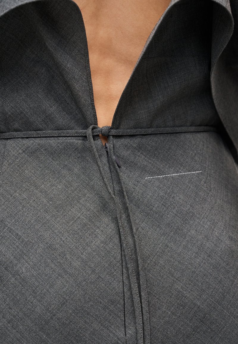 Vêtement gris texturé avec un col muni d'une boucle à l'encolure, un design légèrement ouvert et une couture décorative sur le côté.