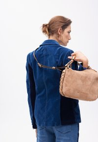 Una donna con un chignon indossa una giacca blu di corduroy e jeans, tenendo una borsa intrecciata beige sulla spalla su uno sfondo bianco.
