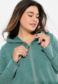 Grüne Zip-Hoodie aus weichem Material, ausgestattet mit Kapuze, genähten Bündchen und einem Reißverschluss mit dekorativem Zugband.