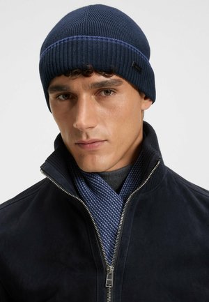 Bonnet tricoté marine avec une texture côtelée et une bande bleu clair contrastante. Porté avec une veste sombre et une écharpe bleue texturée.