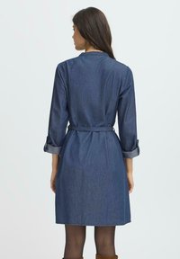 Kvinna med l�ång mörk hår iförd en jeansklänning i knälängd med uppkavlade ärmar och ett midjebälte, stående vänd bort från kameran mot en vit bakgrund.