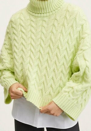 Pull col roulé vert clair en mailles torsadées porté par-dessus une chemise blanche, montré de la poitrine aux hanches avec les mains ajustant l'ourlet.