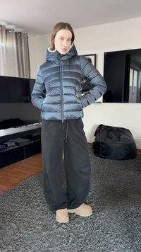 Musta toppatakki hupulla, tikattu kuvio, vetoketjusuljenta, päällä väljien mustien housujen ja beigen väristen jalkineiden kanssa. Sisätilassa maton päällä.