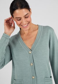 Cardigan verde chiaro con scollo a V, quattro bottoni dorati e due tasche frontali. Tessuto testurizzato con polsini e colletto a costine.