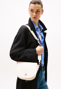 Borsa a tracolla bianca in pelle con patta curva, accessori in argento e tracolla removibile, abbinata a un cappotto nero e a una camicia a righe blu.