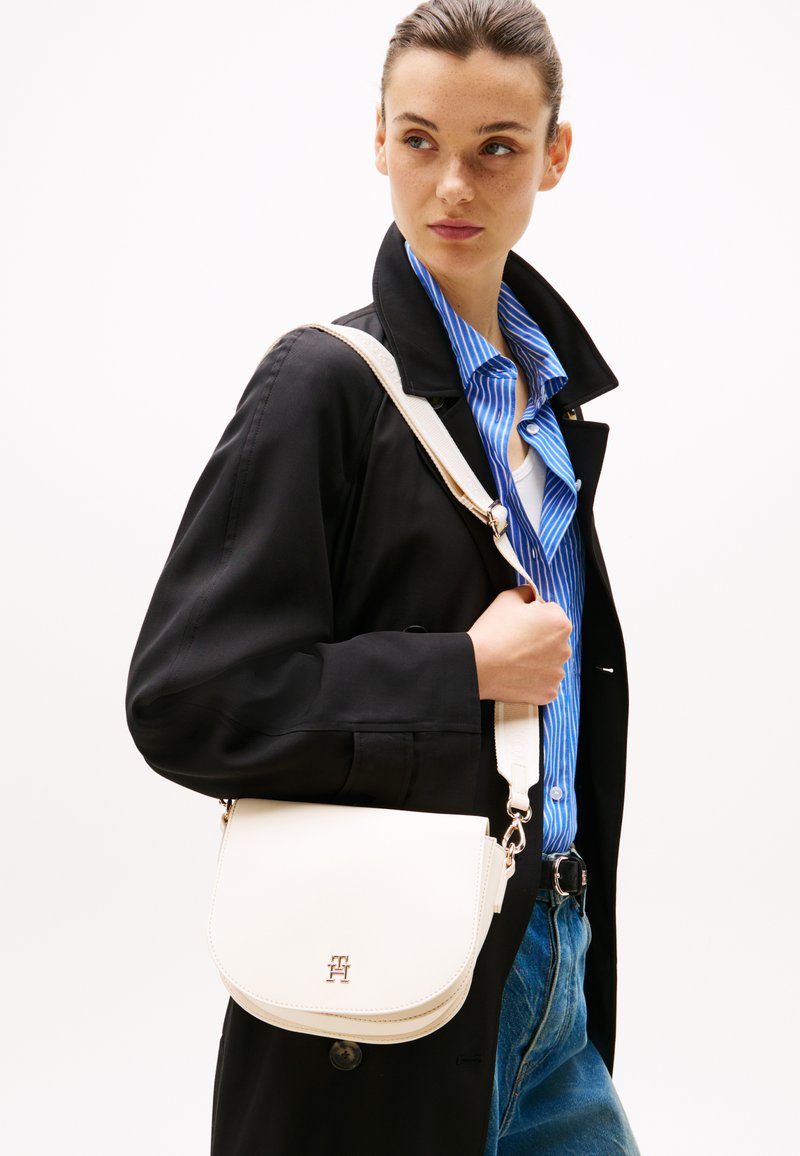 Borsa a tracolla bianca in pelle con patta curva, accessori in argento e tracolla removibile, abbinata a un cappotto nero e a una camicia a righe blu.