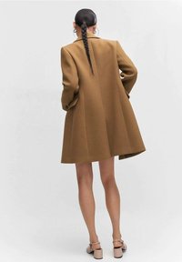 Bruine wollen jas met een gestructureerd, oversized ontwerp. De achterkant heeft een enkele split en het model draagt schoenen in neutrale tonen.