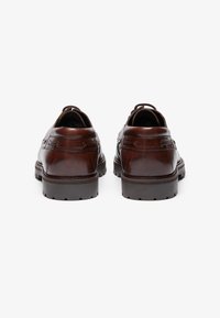 Zapatos de cuero marrón con punta redondeada y diseño curvado, con costuras en contraste, suela de goma y dos lazos decorativos a cada lado.