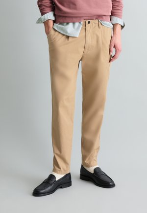 Mann trägt beige Chinos, weiße Socken und schwarze Loafers, steht mit teilweise in die Taschen gesteckten Händen vor einfachem Hintergrund.