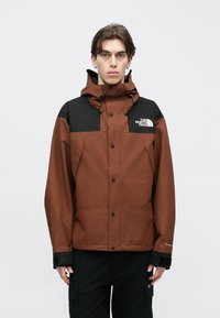 Καφέ και μαύρο αδιάβροχο μπουφάν με κουκούλα, μπροστινό φερμουάρ και κουμπιά με κόπιτσες. Διαθέτει λογότυπο στο στήθος και ρυθμιζόμενα μανίκια. Φαίνονται μαύρο παντελόνι.