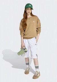 Braunes Sweatshirt, weiße Shorts mit schwarzen Streifen, grüne Mütze, beige Sneakers, hält eine hellgrüne Wasserflasche.