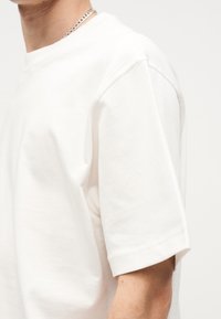 Άνδρας που φοράει ένα απλό λευκό t-shirt με κοντά μανίκια και ασημένιο κολιέ με αλυσίδα σε ουδέτερο φόντο.