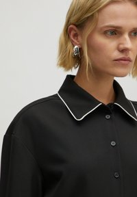 Schwarzfarbiges Hemd mit weißen Paspeln, vorne mit Knöpfen. Das Model hat kurze blonde Haare und trägt silberne Ohrringe.