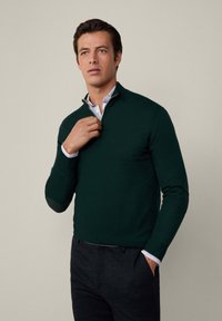 Dunkelgrüner Strickpullover mit einem Viertelreißverschluss, über einem blauen karierten Hemd mit Kragen gestylt, mit Lederbesätzen an den Ellbogen und einer schmalen Passform bei der Hose.