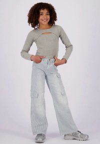 Uśmiechnięta dziewczyna z kręconymi włosami, ubrana w szary, długi top z wycięciem na klatce piersiowej, szerokie spodni w paski oraz srebrne trampki, stoi z rękami w kieszeniach.