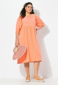 Vestido coral texturizado con escote en V, mangas abullonadas y una silueta acampanada. Combinado con sandalias de plataforma ligeras y un sombrero a rayas.