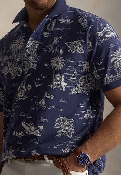 Πόλο μπλουζάκι ναυτικού μπλε με λευκά μοτίβα τροπικού θέματος, που απεικονίζουν ιστιοφόρα, φοίνικες και μια φιγούρα που χαλαρώνει. Φαίνεται ένα καφέ δερμάτινο ρολόι.