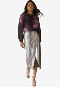 Metallisch glänzender silberner Rock mit Wickeldesign, kombiniert mit einem schwarzen Oberteil, das pinke federartige Muster aufweist. Das Model trägt transparente Riemchen-Heels.