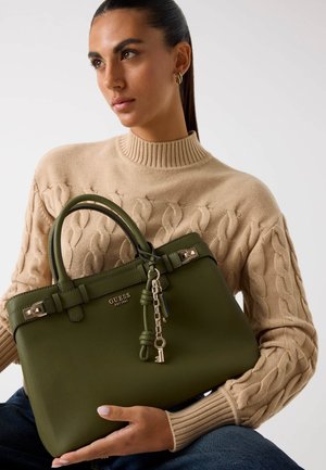 Bolso de mano texturizado verde con doble asa, acentos de hardware plateado y un llavero. La modelo sostiene el bolso mientras lleva un suéter de punto beige.