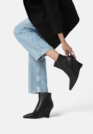 Schwarze Leder-Stiefeletten mit spitzen Zehen und Keilabsätzen, ausgestattet mit einem Seitenreißverschluss. Kombiniert mit hellblauen Jeans.