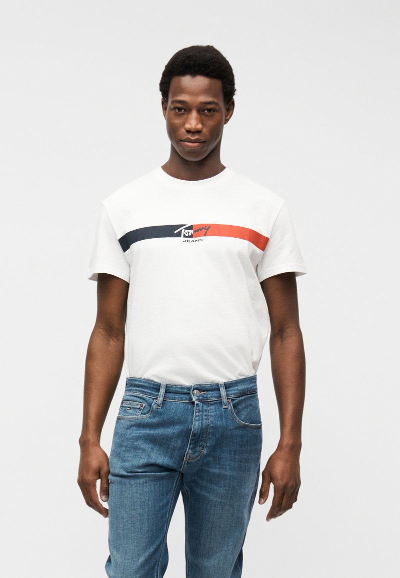 Άνδρας που φοράει λευκό t-shirt Tommy Jeans με ναυτικό και κόκκινο ρίγες και μπλε τζιν, στέκεται μπροστά σε ένα απλό ανοιχτό φόντο.