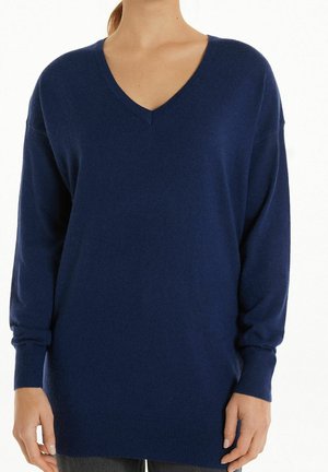 Femme portant un pull bleu marine à col en V, manches longues, avec poignets et bas côtelés.