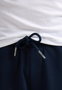 Bílý bavlněný tričko vystavené nad tmavě modrými tepláky. Námořnický šňůrkový pas je svázán s konci se stříbrnými špičkami.