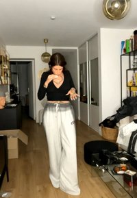 Femme en haut noir et pantalon de jogging gris debout dans un salon, regardant vers le bas et tenant un objet près de sa bouche.