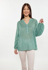Blusa leggera verde menta a maniche lunghe con colletto alla cinese, chiusura a bottoni, polsini elasticizzati e tessuto testurizzato. Abbinata a pantaloni chiari.