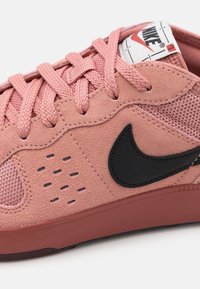 Rožinės zomšinės sportinės bateliai su tinklo ir gumos akcentais, su juodu Nike logotipu, apvaliu nosiu ir lygiais raišteliais. Tekstūra atrodo minkšta ir lanksti.