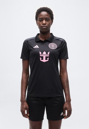 Conjunto deportivo negro con logotipos rosas, llevado por una persona de pie contra un fondo blanco liso, mirando hacia adelante con expresión neutral.