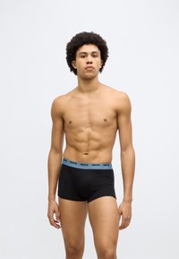 Cuecas boxer pretas com uma cintura elástica azul rotulada com "HUGO." O modelo tem um físico tonificado e um tom de pele natural.