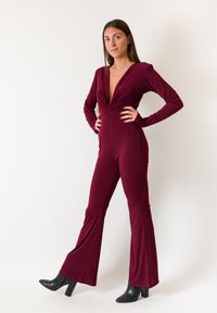 Tuta bordeaux a maniche lunghe con scollo a V profondo e corpetto aderente, caratterizzata da pantaloni svasati. Realizzata in tessuto liscio, abbinata a stivali neri con tacco.