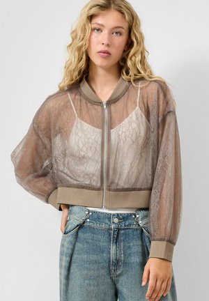 Jeune femme aux longs cheveux blonds bouclés portant une veste bomber en dentelle taupe transparente sur un caraco beige et un jean bleu taille haute.