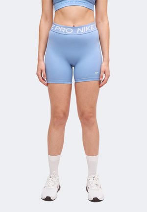Άτομο που φοράει αθλητικό σορτς Nike Pro ανοιχτού γαλάζιου χρώματος με ψηλή μέση, αθλητικό μπουστάκι στο ίδιο χρώμα, λευκές κάλτσες και λευκά παπούτσια για τρέξιμο, στέκεται πάνω σε μονόχρωμο φόντο.