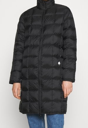 Personne portant un long manteau matelassé noir avec fermeture éclair à l'avant, debout devant un fond clair uni.