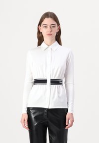 Camicia bianca a maniche lunghe con colletto e cintura elastica nera in vita. Tessuto morbido, design aderente e chiusura con bottoni sul davanti.