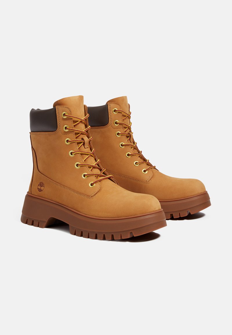 Stivali ankle in suede marrone con lacci gialli, occhielli dorati, colletto imbottito marrone e una suola in gomma spessa e testurizzata. Logo Timberland sul lato.