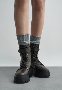 Jambes portant des chaussettes en tricot grises et des bottes de combat à lacets marron foncé avec des semelles épaisses noires, se tenant sur un sol gris contre un fond uni.