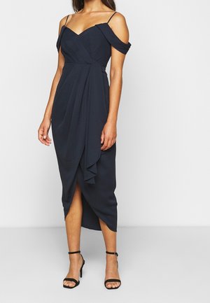 Kobieta w czarnej, midi sukience na ramiączkach, odsłaniającej ramiona, z rozcięciem z przodu, oraz czarne szpilki na kostkę z paskiem.