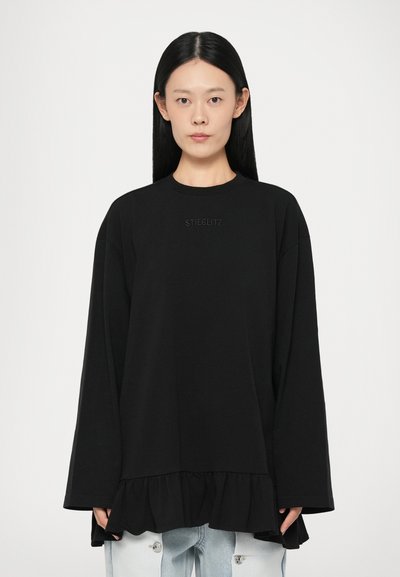 Black long-sleeve top made of cotton, featuring a ruffled hem, round neckline, and subtle "STIEGLITZ" text at the chest.
