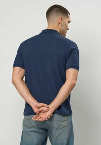 Egy férfi sötétkék rövid ujjú pólóingben és kék farmerben áll, kezeit a háta mögött összekulcsolva, egy sima világos fal felé nézve.