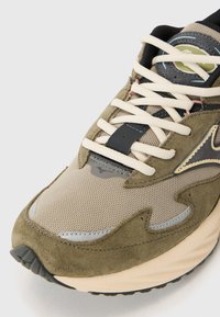 Sneaker with a mix of olive green suede and beige mesh upper, cream laces, and a black rubber sole. Curved design and subtle accents.
