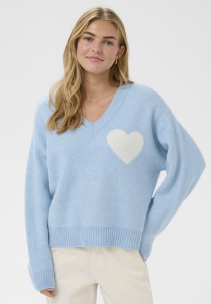 Junge Frau mit langen blonden Haaren, die einen hellblauen Pullover mit V-Ausschnitt und einem weißen Herz auf der Brust sowie cremefarbene Hosen trägt.