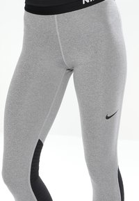 Leggings atléticos grises hechos de una tela elástica con paneles laterales negros. Presentan una cinturilla negra con el logotipo "Nike" y un pequeño swoosh negro.