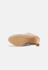 Chaussure à talons hauts pour femme en daim beige, couchée sur le côté, montrant une semelle lisse et un talon effilé sur fond blanc.